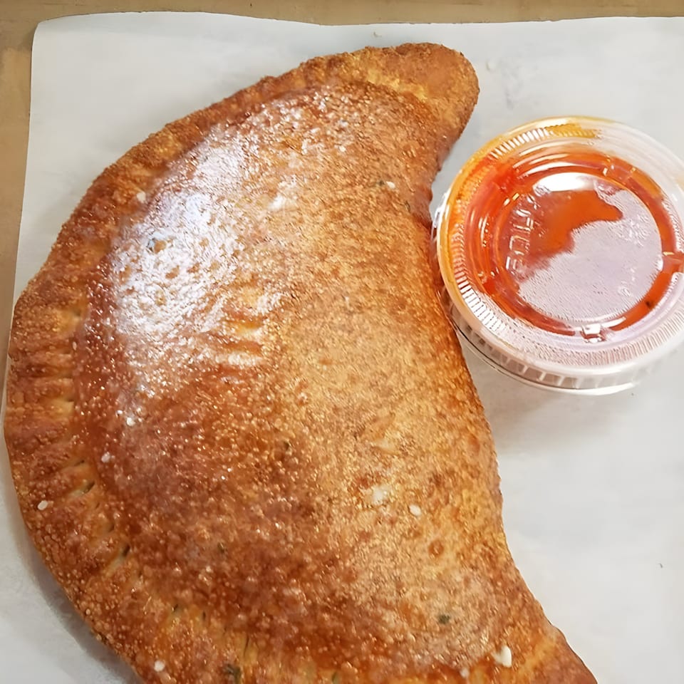 Golden-brown calzone from Pizza Roma, sprinkled with salt, next to a small container of red sauce.